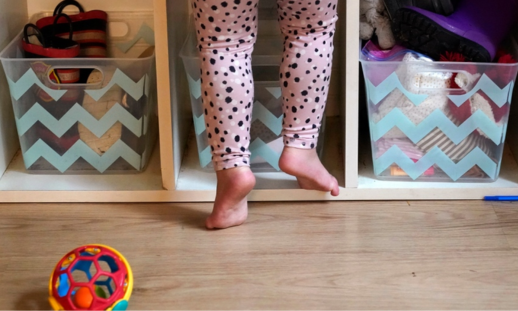 image shows a baby's legs, leaning against a bookshelf with toys. 