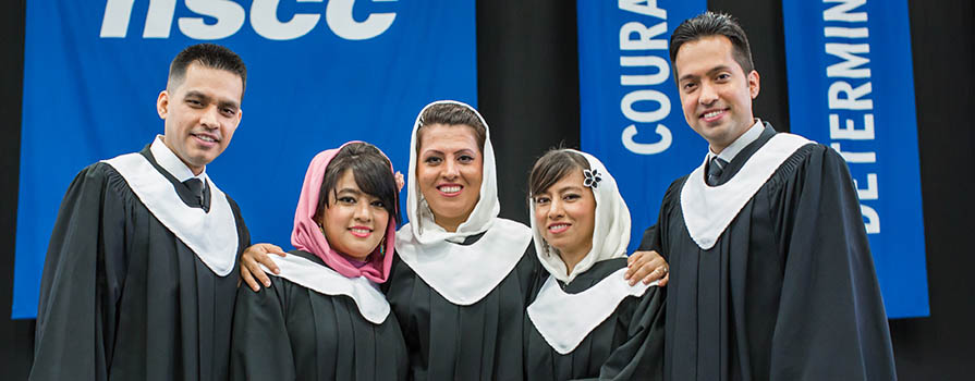 Five people stand shoulder to shoulder in black graduation gowns and white stoles. Three women in the centre wear hijabs and the men at each end wear collared shirts and ties. In the background, fabric signs read N S C C, courage and determined.