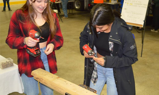 Two female students use a drill on a piece of wood as part of a live demonstration at a recent career fair.