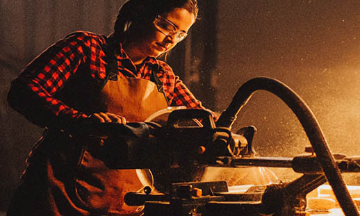A woman in safety glasses and a plaid shirt works with a reciprocating saw.