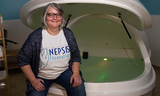 A woman sits on the edge of a large, white flotation pod that is filled with water. She is wearing a sweater over a t-shirt with nepsis written on it. She is smiling. 