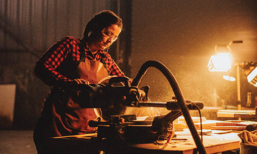 A woman wearing a buffalo plaid shirt, leather apron and safety glasses, cuts wood with a power saw. Sawdust can be seen in the air. There is a light shining in the background.