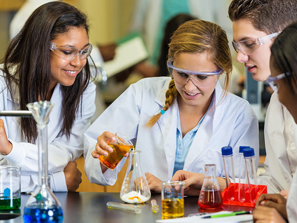 Four students, wearing white lab coats and safety glasses, conduct an experiment in a science lab.