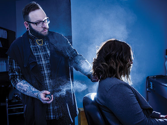 A stylist sprays hairspray on a client's freshly-cut hair in a salon setting.