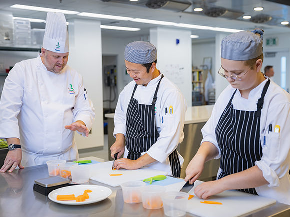 Two students wearing chef hats and aprons work alongside their chef instructor seen wearing a chef hat and uniform whites.