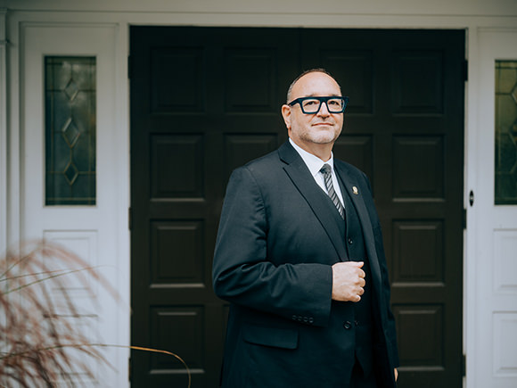 A funeral director stands in a suit in front of a funeral home.