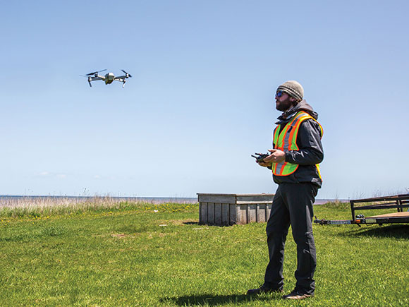 A man in a safety vest stands in a field while operating a drone.