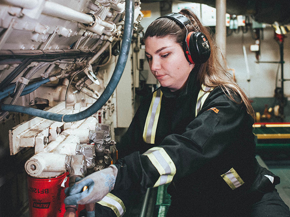 A woman in protective gear works on engine room equipment.