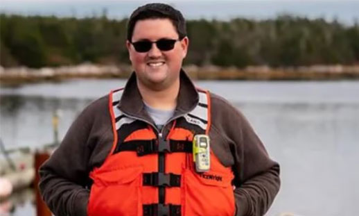 Matthew Duffy is shown. He is wearing a life jacket and standing with his back to a body of water.