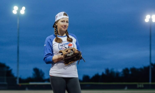 A woman wearing a Toronto Blue Jays shirt, a baseball cap and baseball glove stands in a baseball field under bright stadium lights.