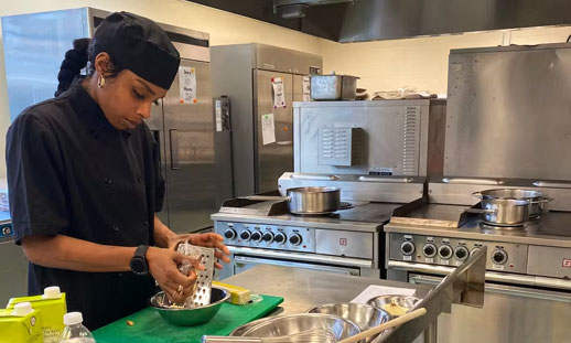 A woman is shown in a professional kitchen, wearing an apron and chef's hat as she focuses, preparing food.