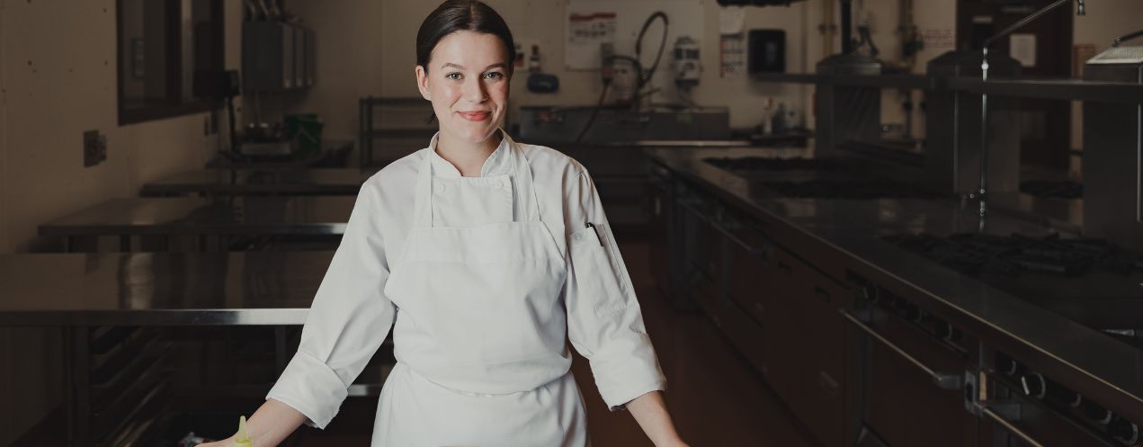 Olivia Sewell, a culinary management student, stands in a chef jacket and apron in an industrial kitchen