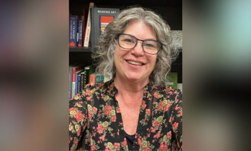Pam Eakin sitting in front of a bookshelf filled with books, wearing a black floral-patterned top.