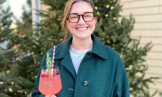 A woman holding a red drink, smiling in front of a spruce tree with lights on it.