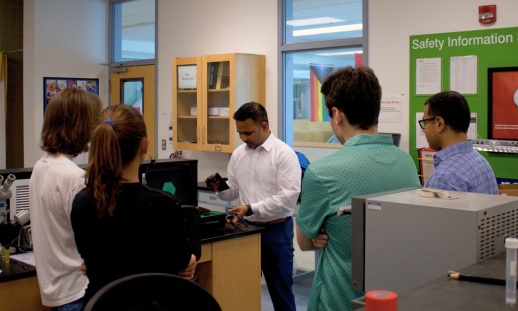 A group of four young adults standing around a table, observing a man in a lab setting as he demonstrates a water system.