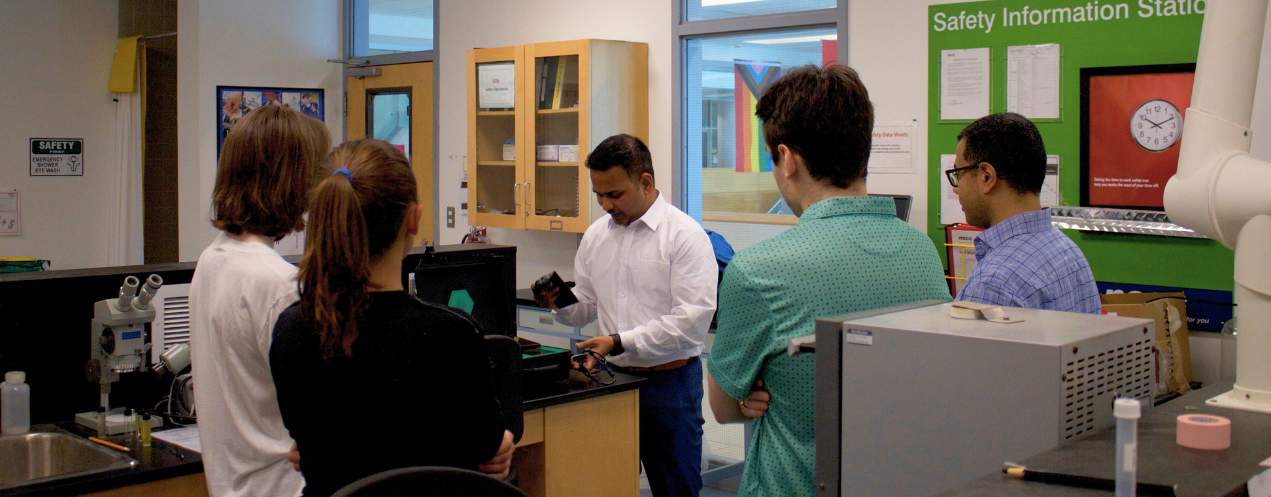 A group of four young adults standing around a table, observing a man in a lab setting as he demonstrates a water system.