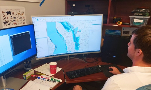 Researcher works on two monitors looking at maps and data.