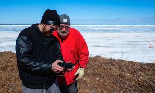 Two men standing on the coast looking at a piece of technology with ice in the background.