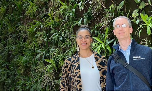 A woman and man standing together smiling in front of a plant wall. 