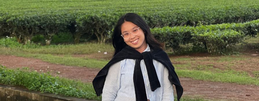 A young woman standing in a green field smiling with a sweater wrapped around her shoulders.