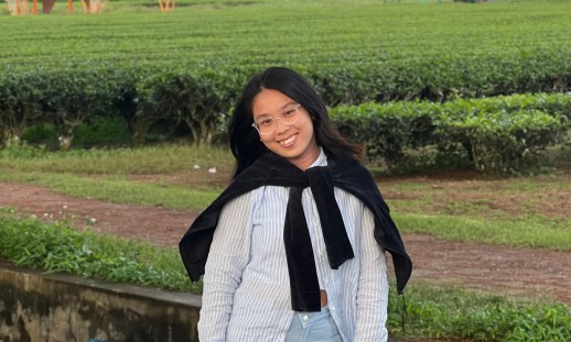 A young woman standing in a green field smiling with a sweater wrapped around her shoulders.