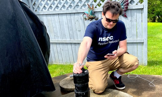 A man wearing an NSCC Applied Research t-shirt is leaning over a residential well, seemingly tracking something on his phone with a piece of equipment.