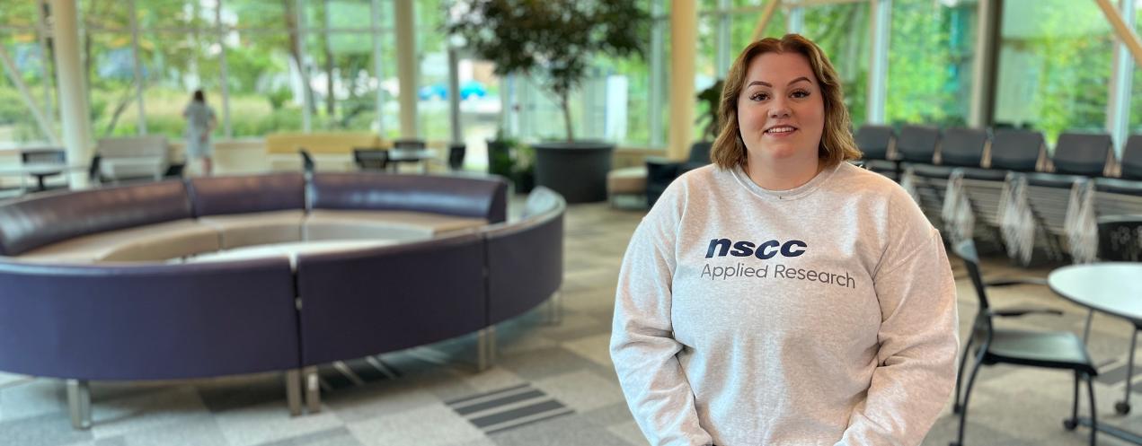 A young woman wearing an NSCC Applied Research sweatshirt standing in the college lobby smiling.