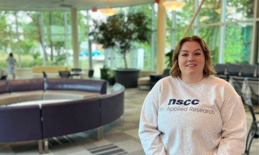 A young woman wearing an NSCC Applied Research sweatshirt standing in the college lobby smiling. 