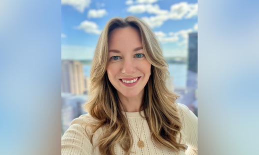 Photo of Niecole Killawee in front of a window with the background showing a bright cityscape with tall buildings and a body of water under a blue sky with scattered clouds.