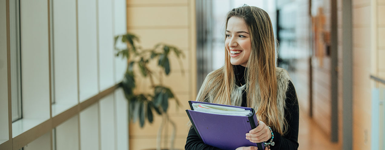 A student carries a binder and smiles as she walks through an NSCC campus building.