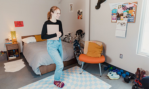 A student standing, holding a laptop while in campus housing