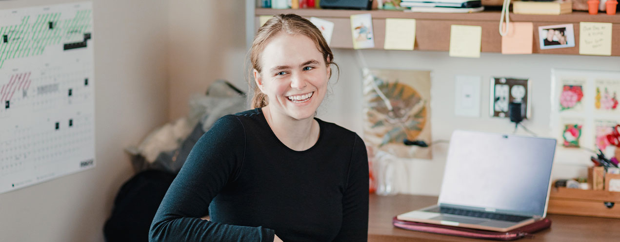 A student smiling, while sitting at a desk with a computer in a background.