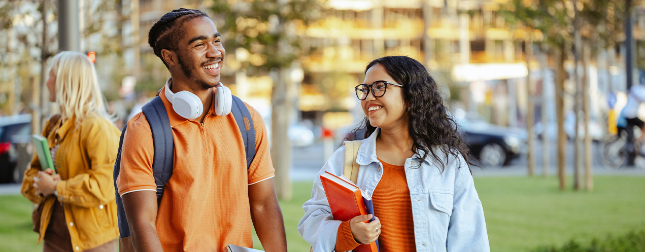 Two students walking along side one another, talking and smiling, one holding a book and one wearing headphones