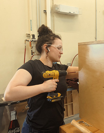 Cabinetmaking student, Mackenzie, uses a power drill to screw together a kitchen cabinet project.