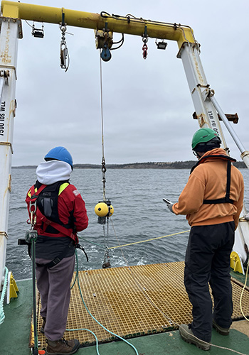 Emma and instructor Stephane stand toward the back of a ship, lowering a device into the water. They are wearing protective safety gear.