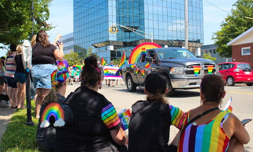 Parade attendees are sitting on the side of the road while pride parade floats drive by in Cape Breton.