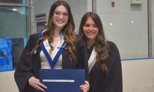 Two students stand together holding their diplomas at graduation.