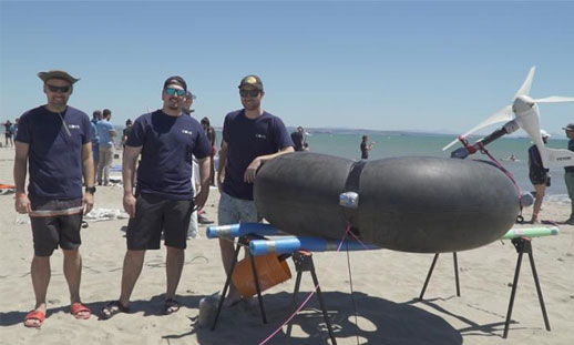 Three students stand together on a beach beside a floating wind turbine.