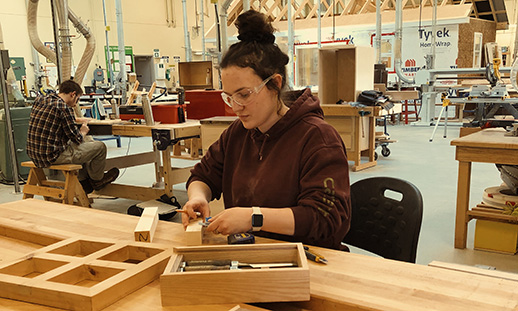 Mackenzie is a Cabinetmaking and Woodworking Techniques student pictured seated at a work table in the Cabinetmaking workshop. She is using a measuring tape to inspect a wood joinery project.