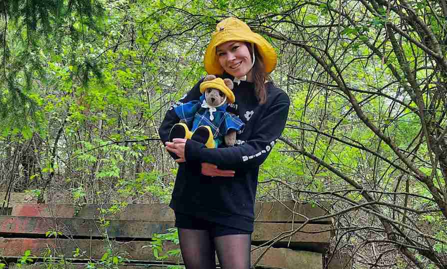 A woman in nature holding a hand-knit stuffed bear dressed in Nova Scotia tartan and a matching yellow sou'wester hat.
