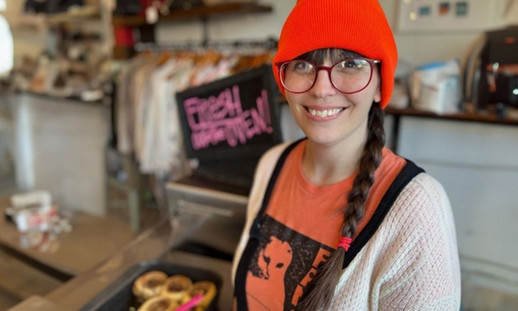 Smiling woman wearing a bright coral toque, standing in front of baked goods inside a trendy shop.