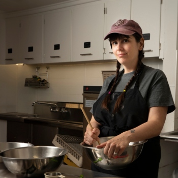 A woman wearing an apron stirring ingredients in a stainless steel bowl, surrounded by other bowls in a kitchen.