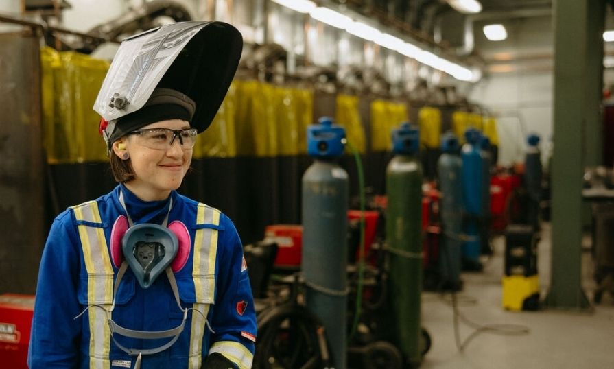 Woman in welding protective clothing with raised face shield, positioned in a metal fabrication shop.