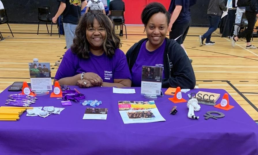 Two women smiling while seated at a purple-themed booth at an event.