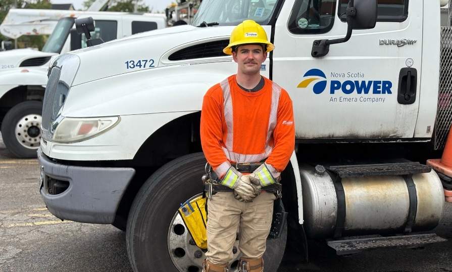 A power line technician trainee wearing a hard hat stands in front of a utility truck during hands-on training.