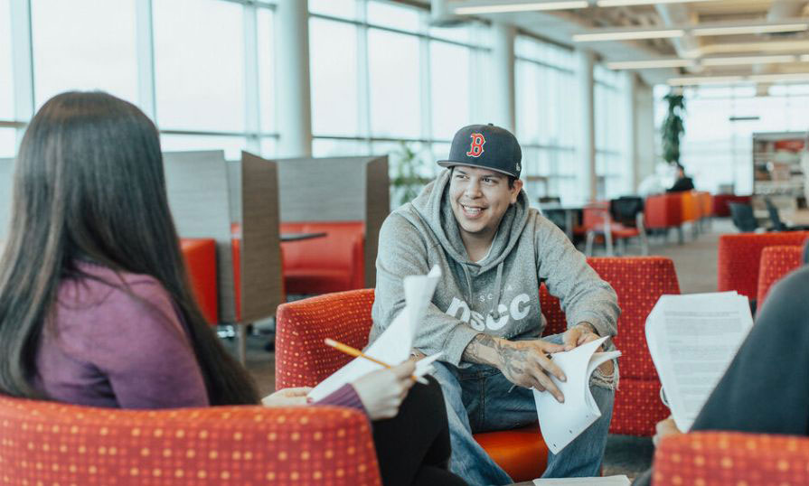 A student wearing an NSCC grey hoodie and a baseball cap talks to two other people shown from behind in a library setting.