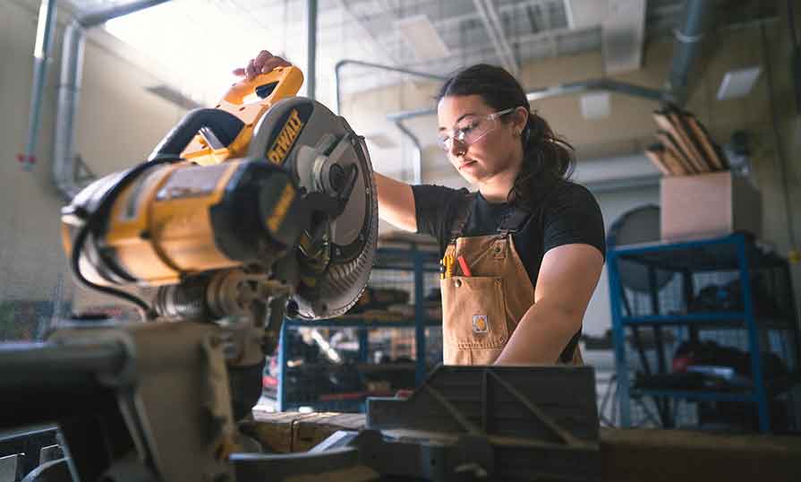 A woman in safety glasses with her hair pulled back is seen working with a huge table saw in a workshop setting.