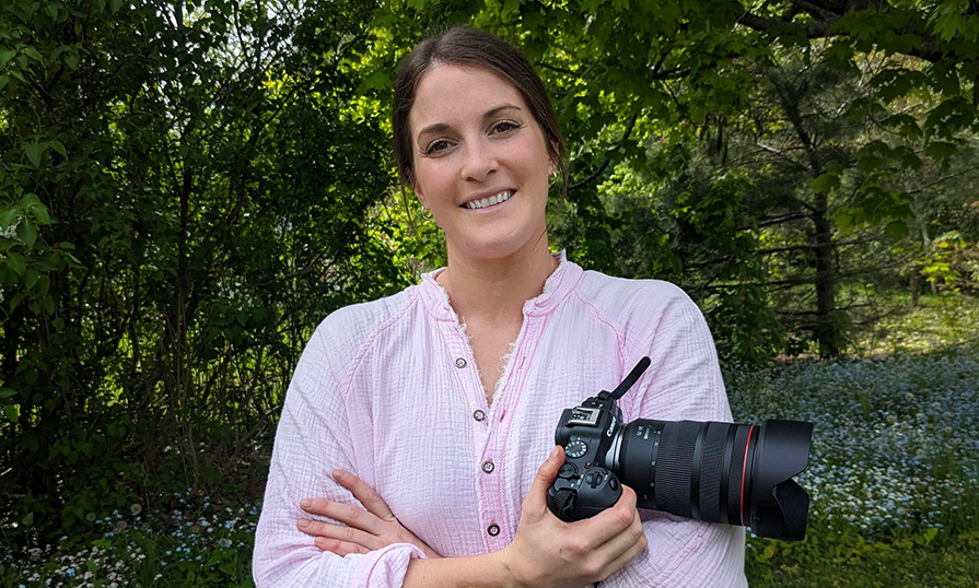 A person standing in front of trees holds a camera while smiling at the camera.