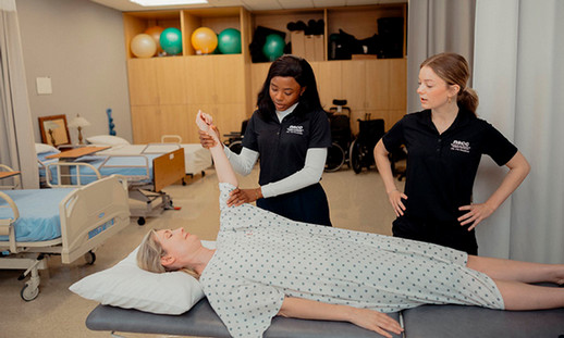 Two students stand in a clinical environment next to a patient on a bed. One student holds the patient's arm.