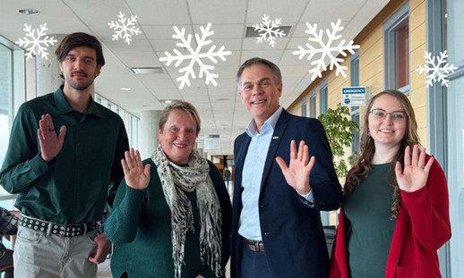 Four people stand facing the camera, smiling and waving. Graphic designed white snowflakes are scattered around the image.